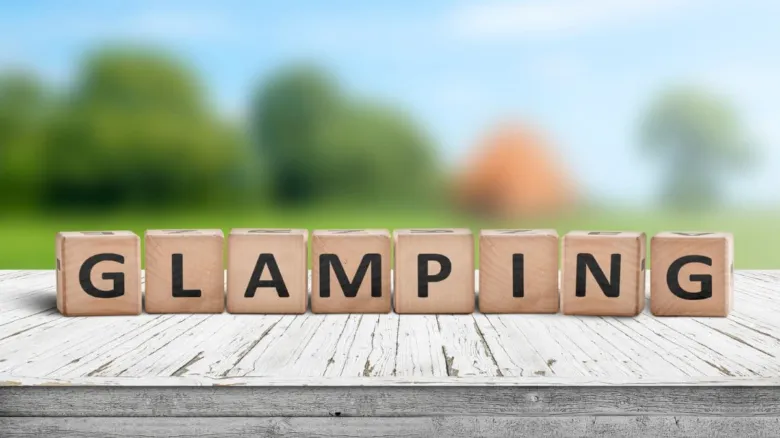 Dřevěné kostky s nápisem GLAMPING na venkovském pozadí.