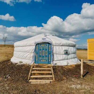 Bílá jurtová glamping chatka na travnatém terénu pod modrou oblohou.