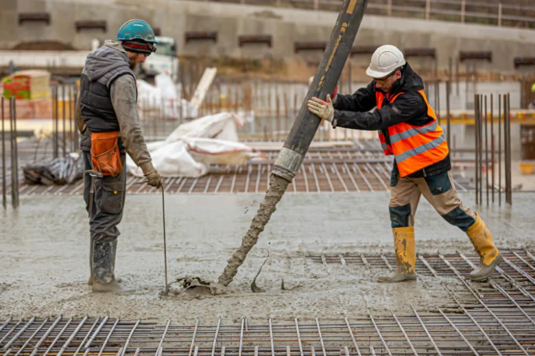 Dva dělníci nalévají beton na armaturu v základové desce.