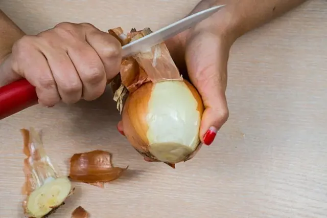 Ruce škrábou slupku z cibule nožem na dřevěném povrchu.