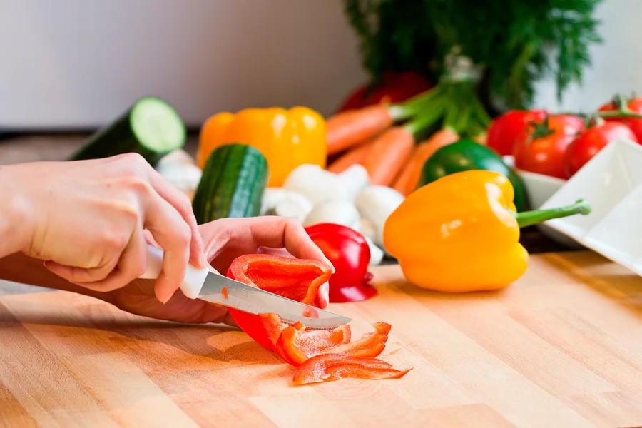 Ruční řezání červené papriky na dřevěném prknu s dalšími zeleninovými ingrediencemi.